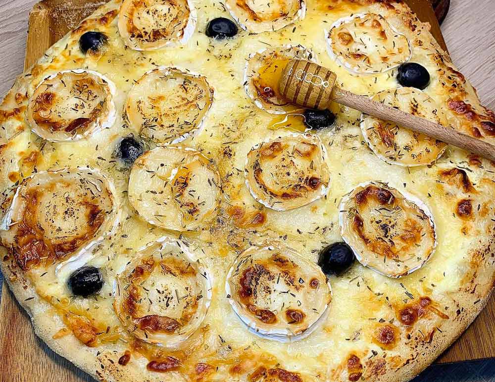 Savourez notre pizza artisanale cuite au feu de bois, garnie de crème, mozzarella fondante, emmental, chèvre, miel et olives. Une recette gourmande et généreuse,