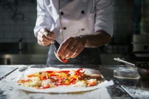 Pizzaiolo en train de garnir une pizza artisanale avec des légumes frais et du fromage dans une cuisine professionnelle – cuisson au feu de bois, savoir-faire traditionnel.